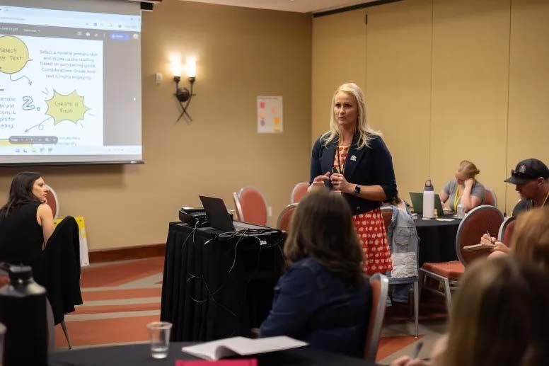 Woman presenting to a group in a conference room, using a projector. Attendees seated at tables.