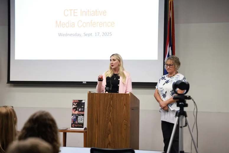 Woman speaking at a podium during a media conference with another woman standing beside a tripod-mounted camera.