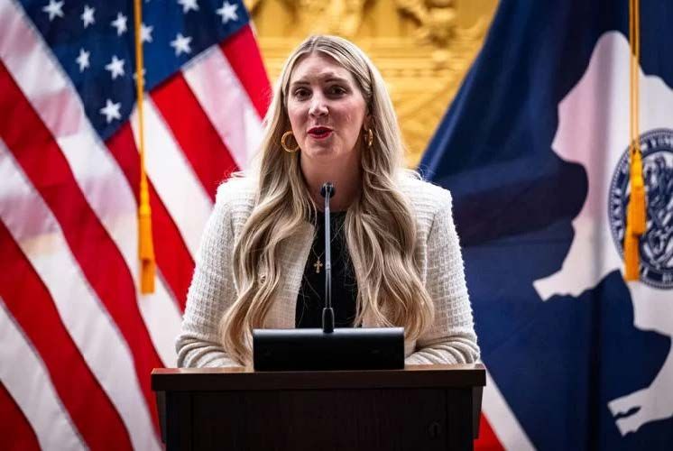 Woman speaking at a podium with US and Wyoming flags in the background. She wears a cream jacket and has blonde hair.