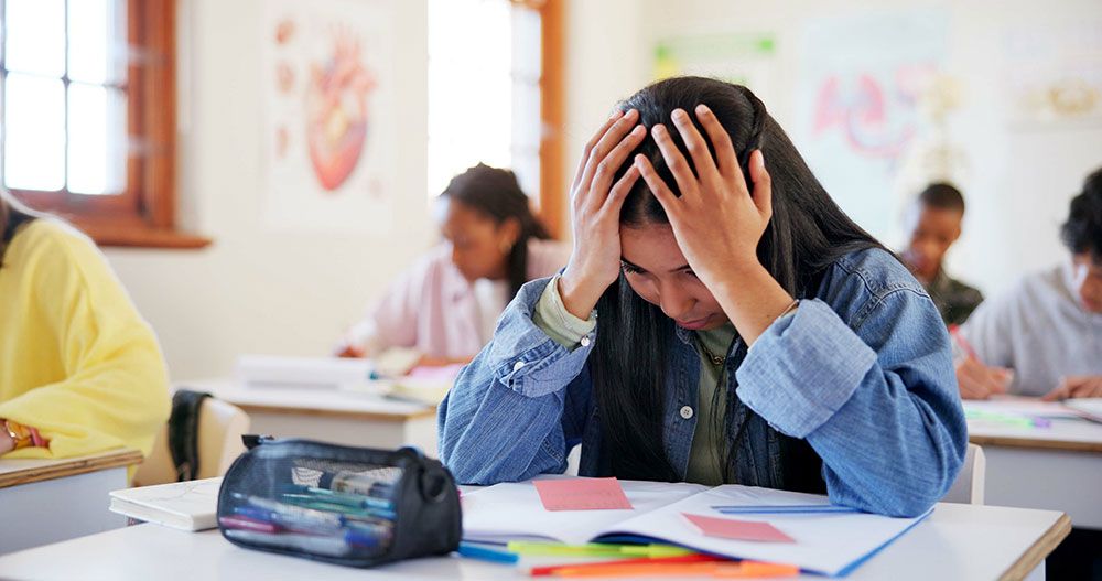 Student in a classroom with hands on head looking stressed over notebook at desk.