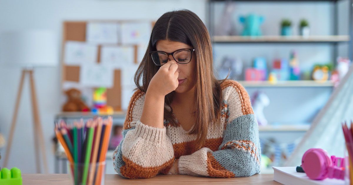 Woman wearing glasses, resting hand on face, looking stressed, sitting at a desk with colored pencils and supplies.