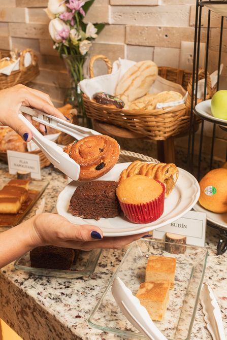 Un par de manos utilizan pinzas blancas para colocar un pastelito en un plato lleno de un brownie, una magdalena y otro pastelito en un bufé.