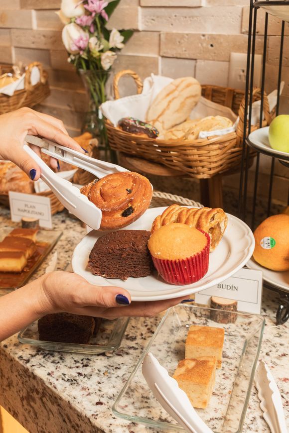 Un par de manos utilizan pinzas blancas para colocar un pastelito en un plato lleno de un brownie, una magdalena y otro pastelito en un bufé.