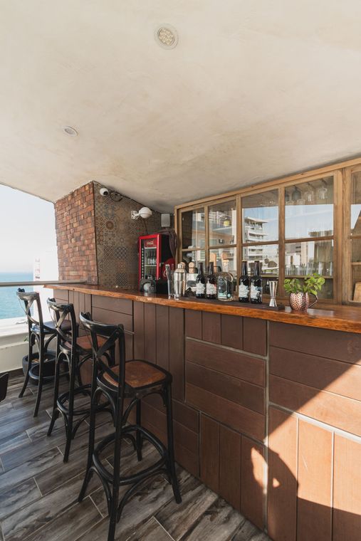 Un bar rústico al aire libre con paneles de madera, sillas altas negras y vistas al mar desde un balcón.