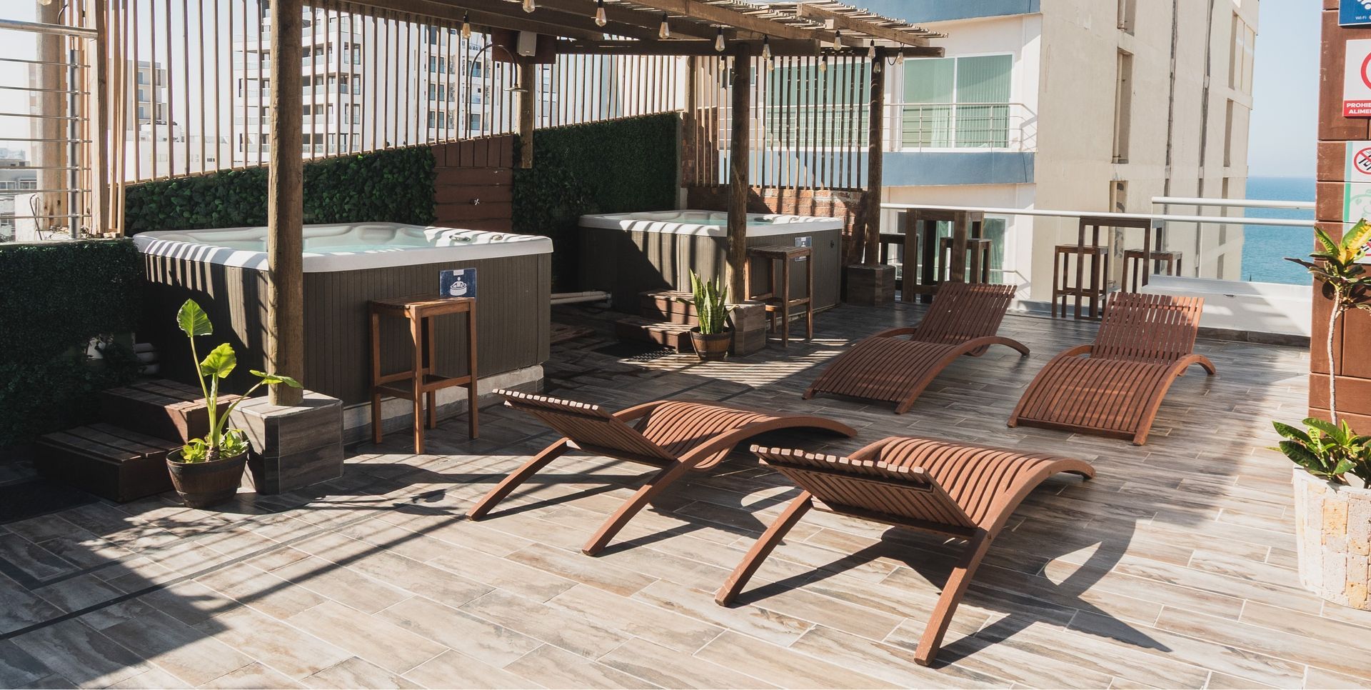 A rooftop deck with two hot tubs, wooden lounge chairs, and an ocean view in the background.