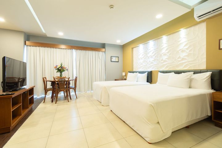 Una habitación de hotel con dos camas blancas, una mesa de comedor de madera con sillas, un televisor y grandes cortinas blancas.