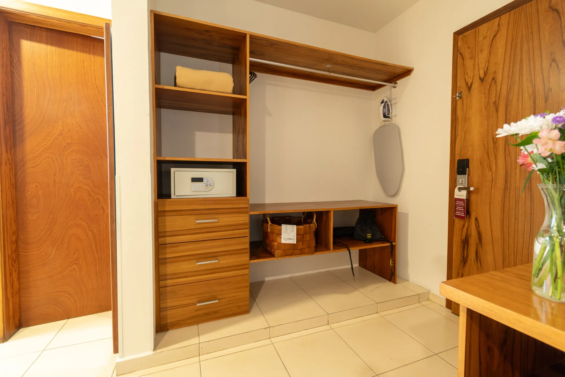 Un mueble de almacenamiento de madera con estantes, cajones, una caja fuerte y un área para colgar ropa con tabla de planchar en una habitación con piso de baldosas.