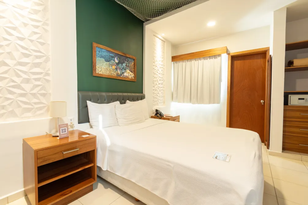 Una habitación de hotel con una cama blanca con almohadas, una pared decorativa de color verde, una mesita de noche de madera y un armario abierto cerca.