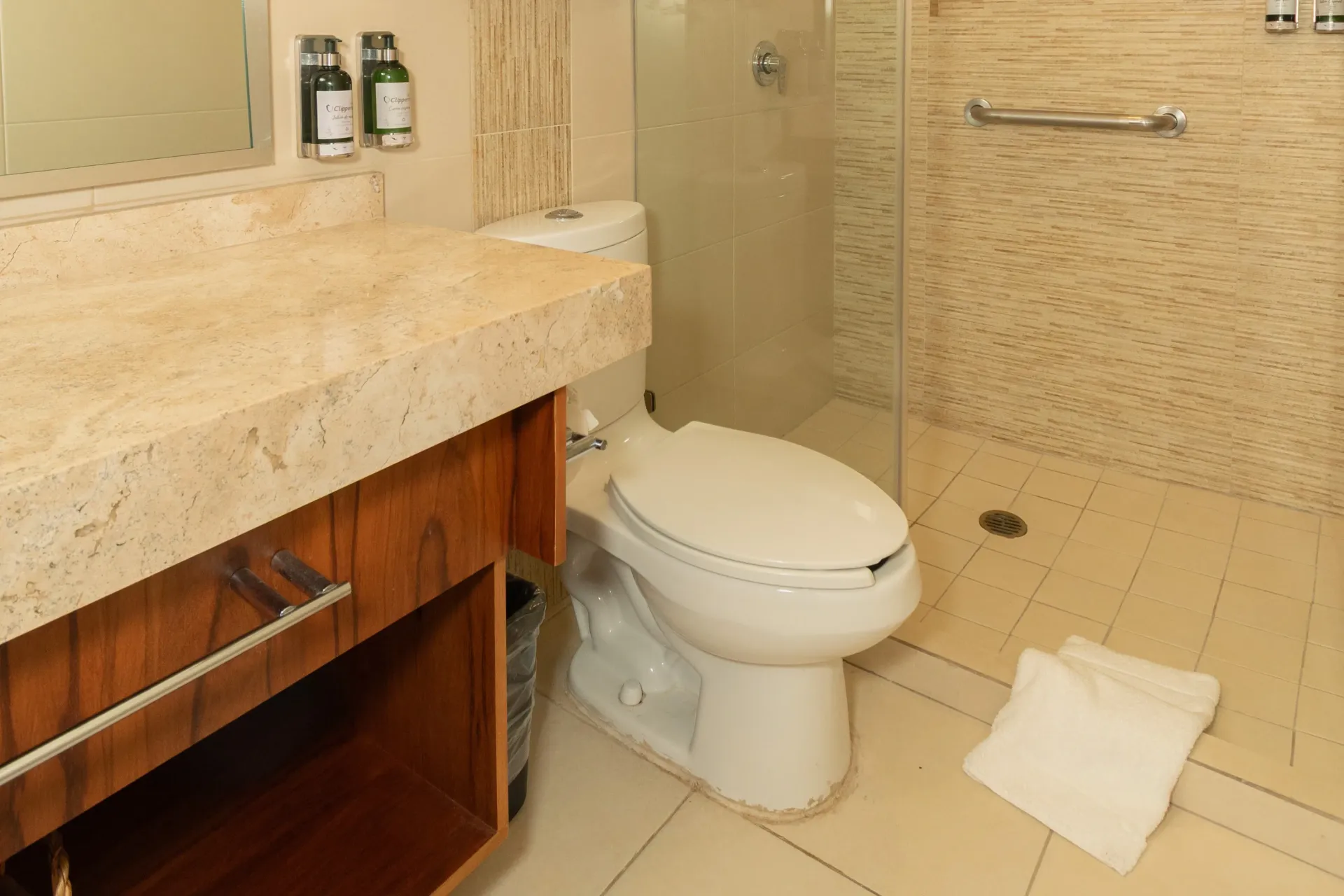Un baño moderno con tocador de piedra beige, armarios de madera, inodoro blanco y ducha con mampara de cristal y barra de apoyo.