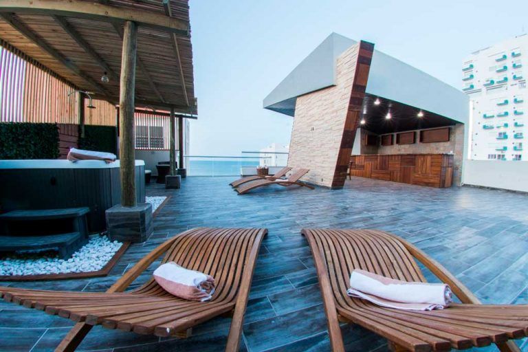 Two wooden lounge chairs are sitting on a rooftop with a hot tub in the background.