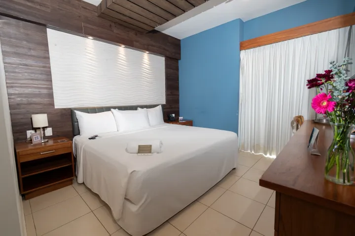 Una habitación de hotel con una cama grande blanca, paneles de madera oscura en las paredes, una pared decorativa azul, suelo de baldosas y un jarrón con flores.