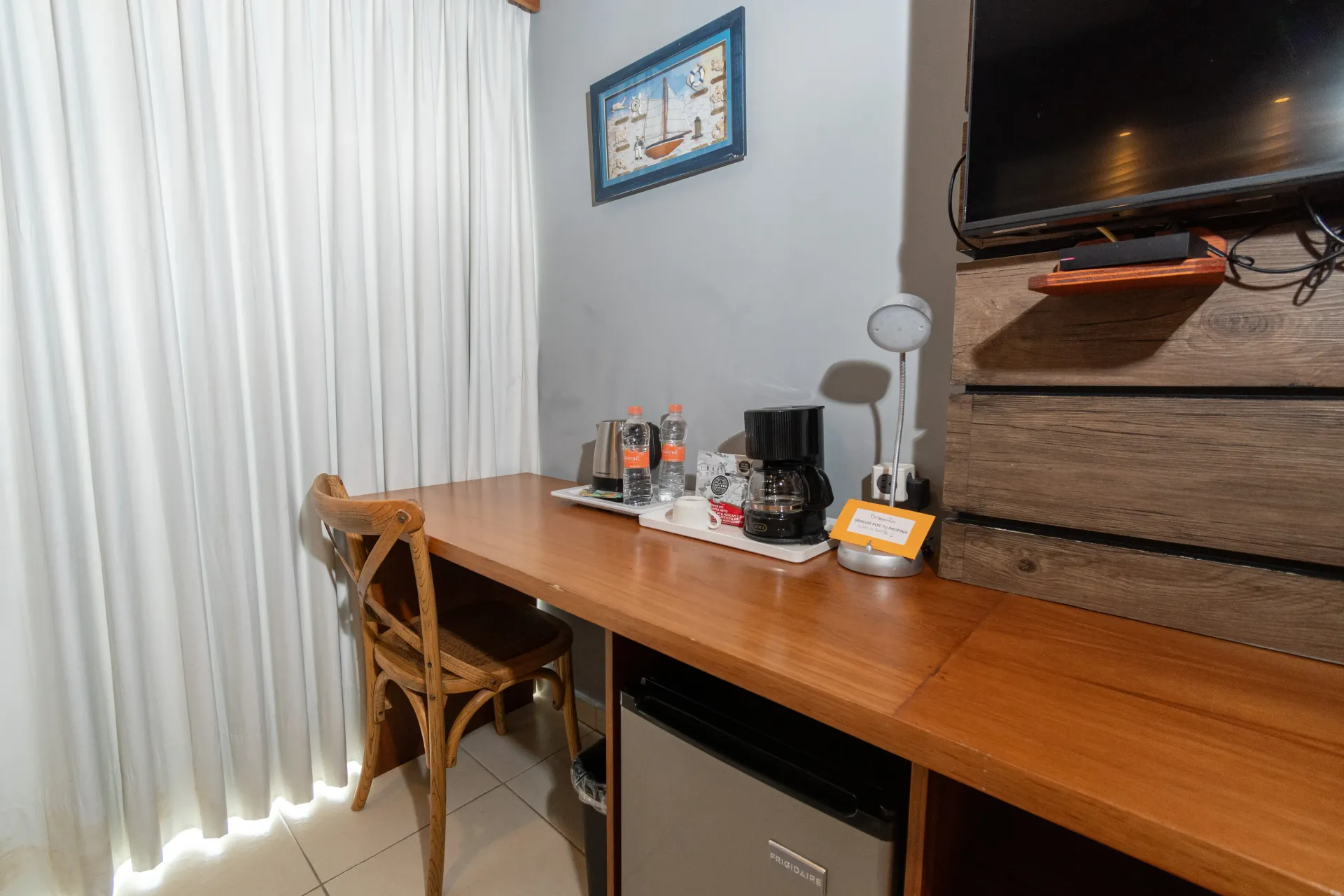 Un escritorio de madera con cafetera y lámpara se encuentra junto a una silla y cortinas blancas, debajo de un televisor y cuadros en la pared.