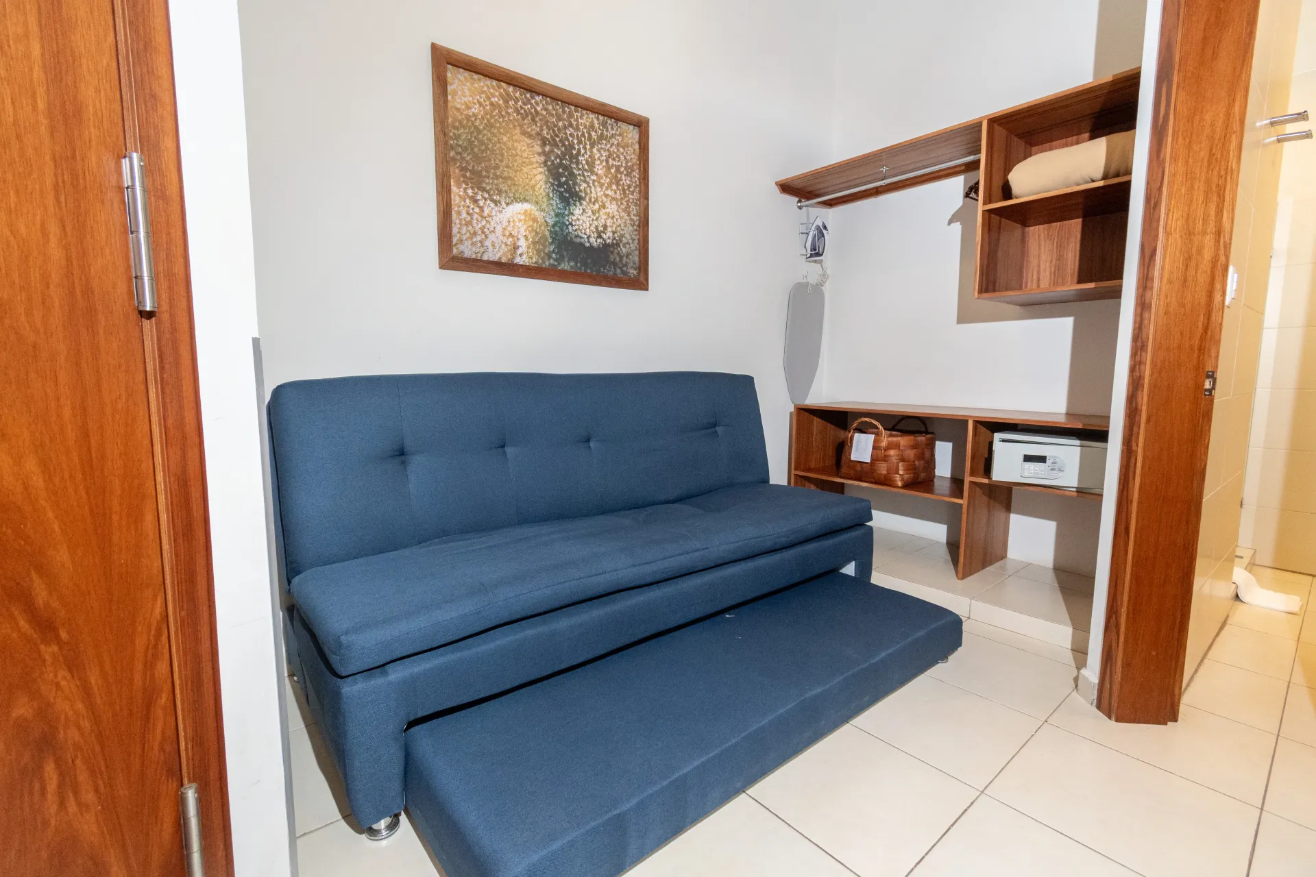 Un sofá cama nido azul se encuentra en una habitación con suelo de baldosas de color claro y estantes de madera montados en la pared.