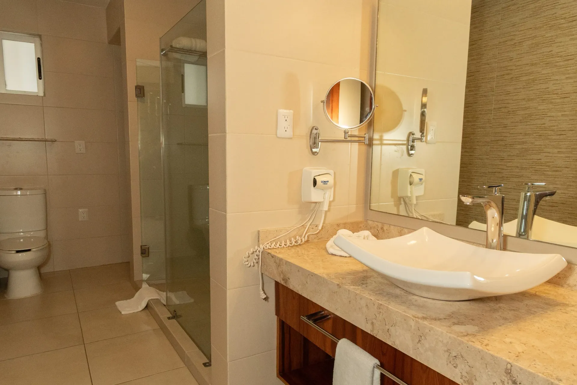 Un moderno baño de hotel con lavabo blanco sobre encimera de piedra color tostado, espejo de pared y ducha de cristal.