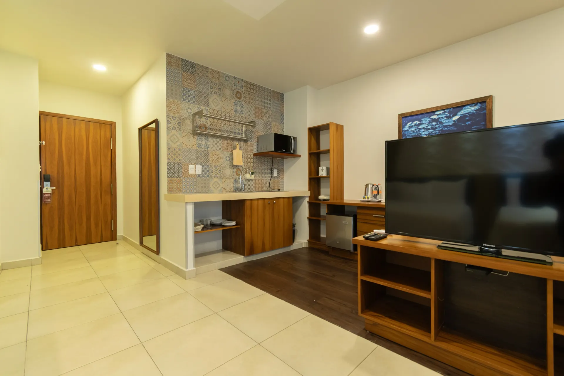 El interior de una habitación de hotel cuenta con una cocina pequeña con salpicadero estampado, armarios de madera, un soporte para la televisión y una puerta de madera.