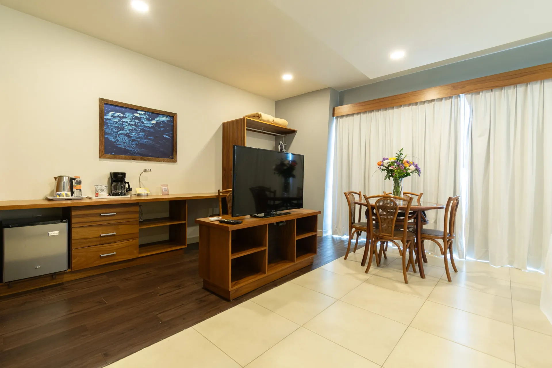 Interior de una habitación de hotel con mueble para televisión, cocina americana con mininevera y una pequeña mesa de comedor con cuatro sillas.