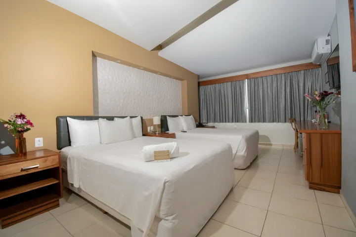 Una habitación de hotel limpia con dos camas blancas, una mesita de noche de madera con flores y ventanales que van del suelo al techo con cortinas grises.