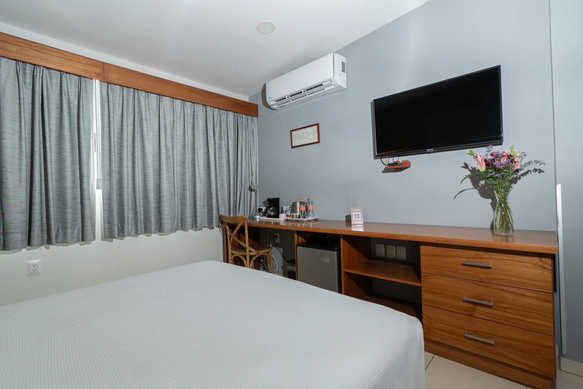 Una habitación de hotel con una cama, cortinas grises, un escritorio de madera con un televisor montado encima y un jarrón con flores.