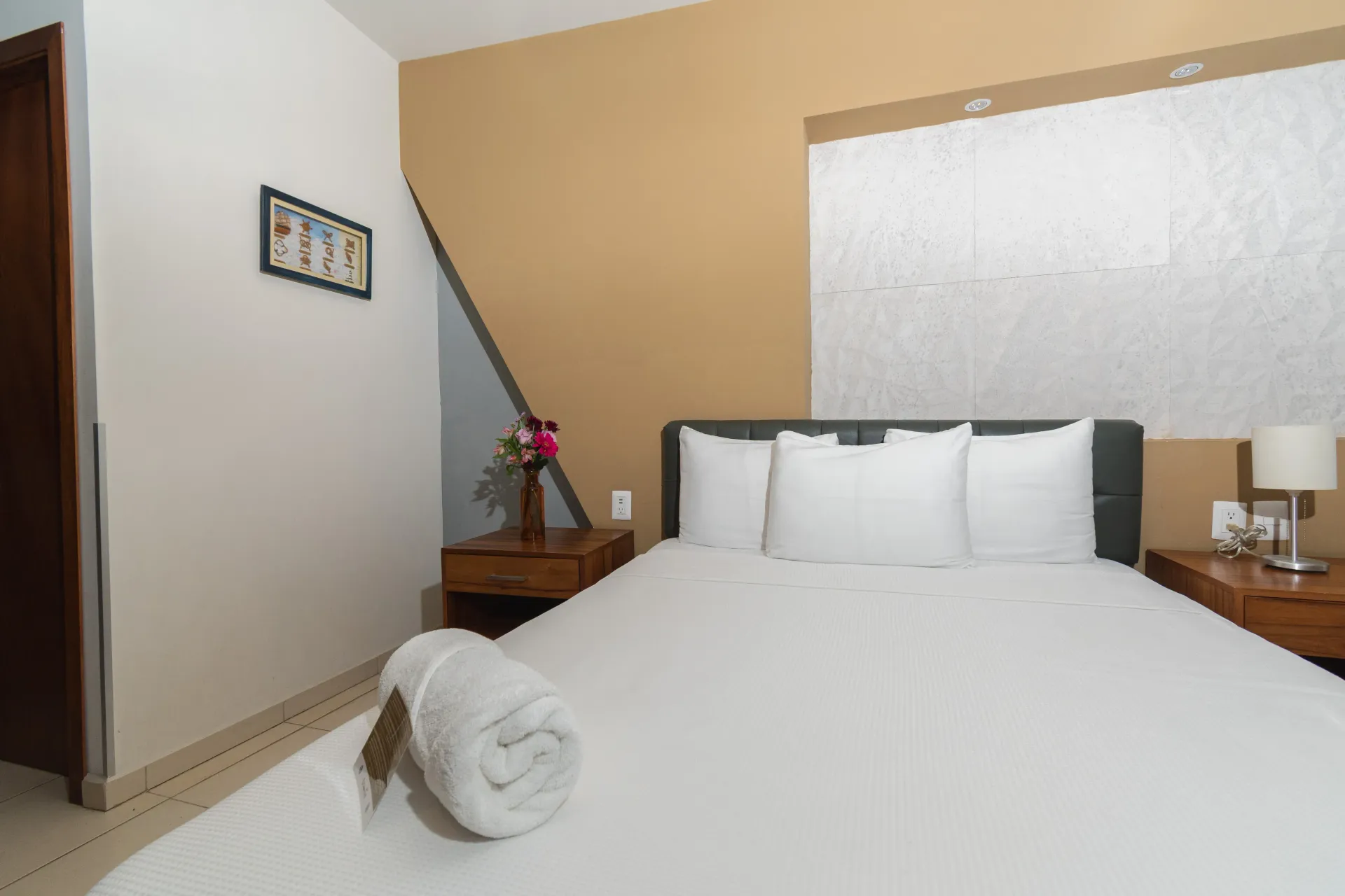 Un dormitorio con una cama con ropa de cama blanca, dos mesitas de noche de madera y una pared decorativa en tonos beige y gris.