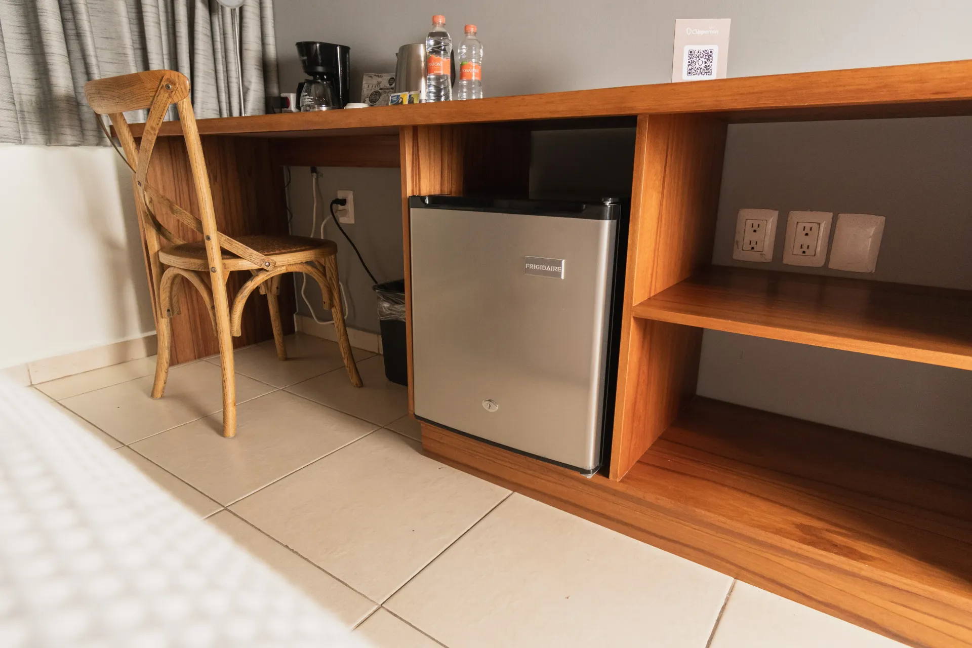 En una habitación de hotel, un escritorio de madera con silla, una cafetera, agua embotellada, un pequeño refrigerador y estantes.