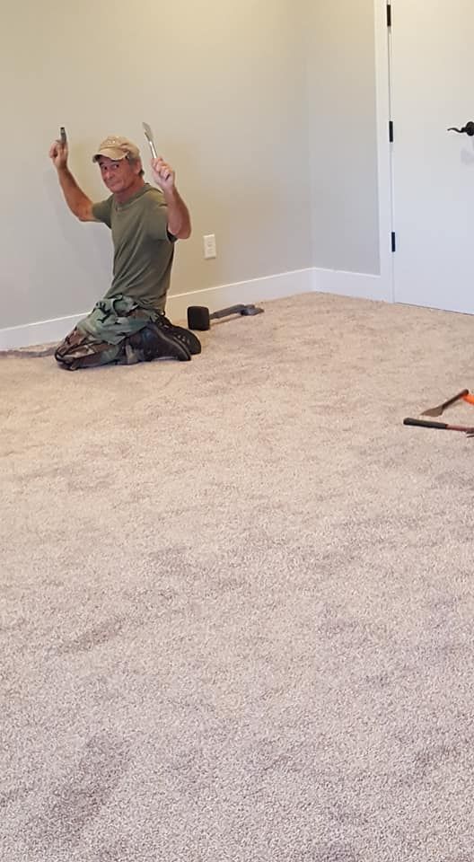 Man kneeling on new carpet, arms raised holding tools. Neutral-colored walls and door in the background.