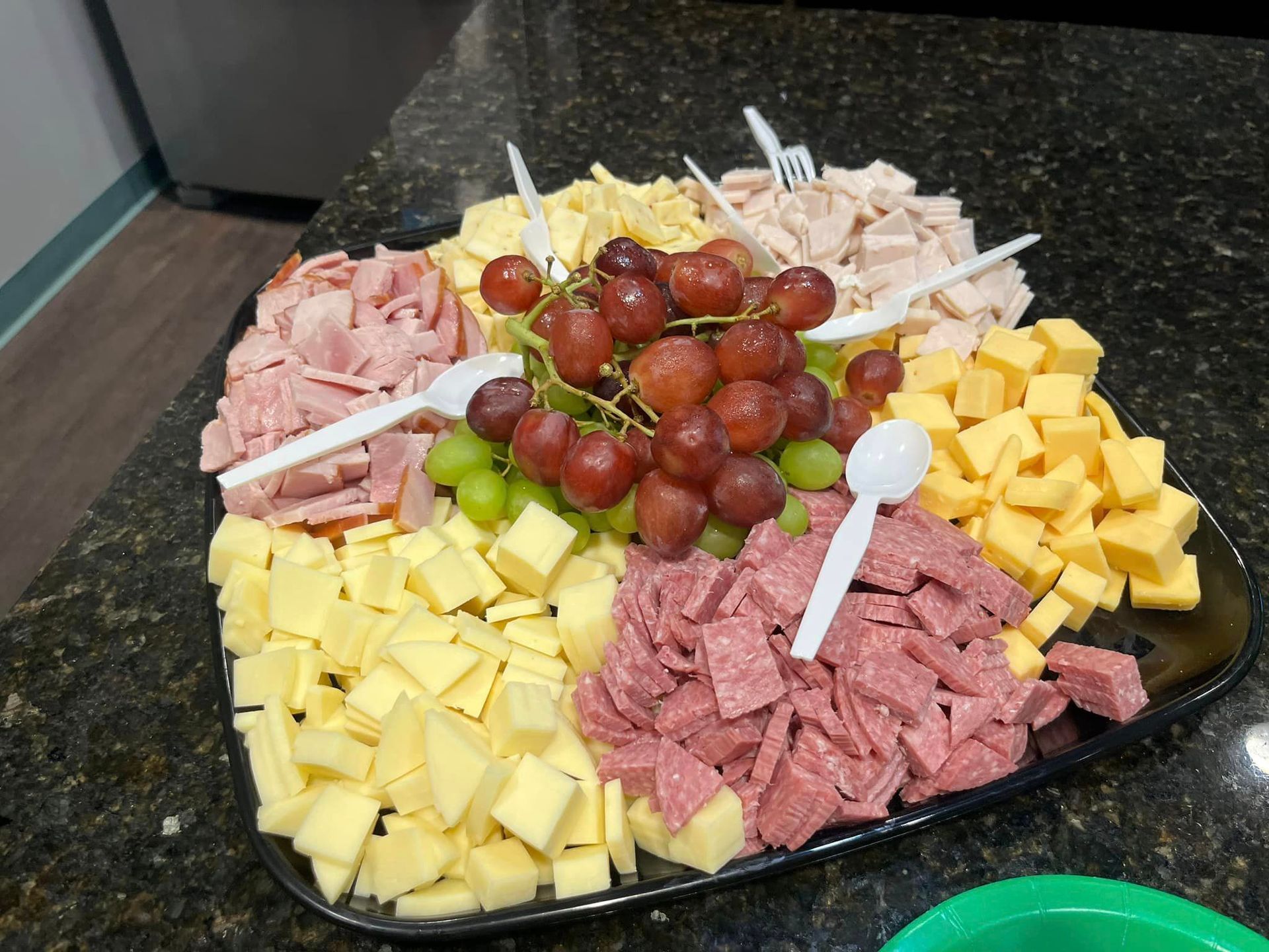 A tray of meats , cheeses , grapes and spoons on a table.