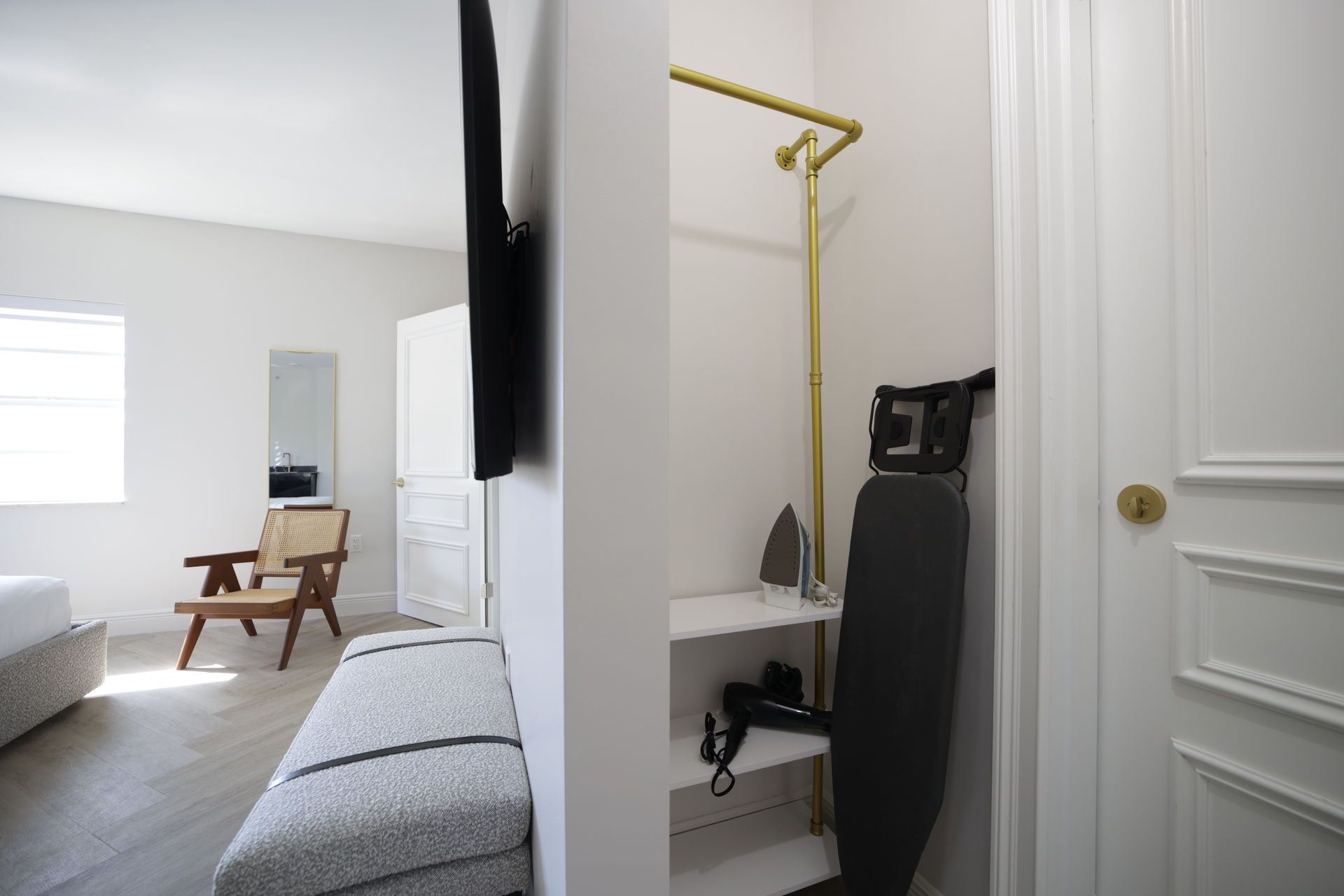 A closet with a hanging rod, ironing board, and shelves. A chair and window are in the adjacent room.