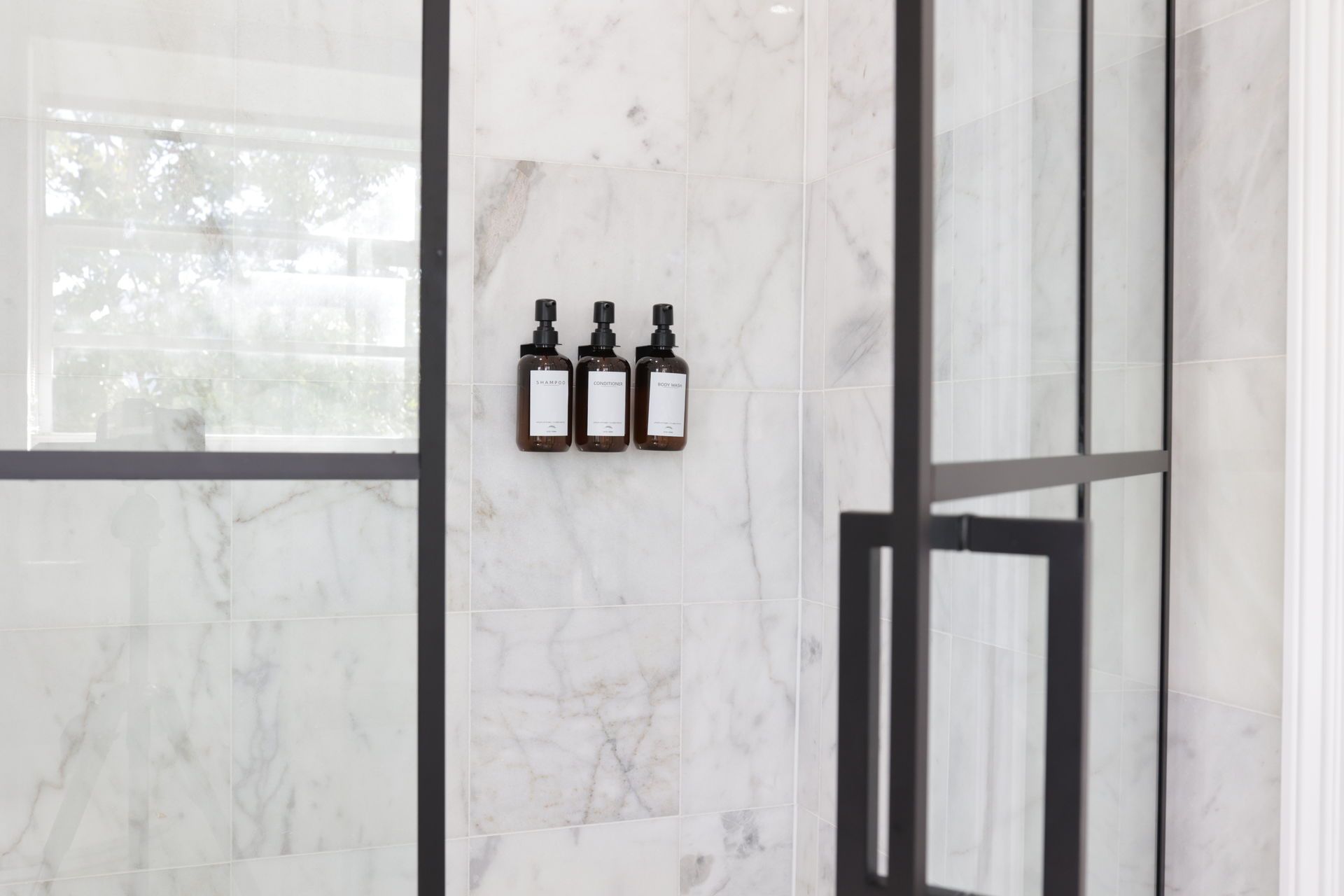 Three dark pump bottles mounted on a marble-tiled shower wall, behind a black-framed glass door.