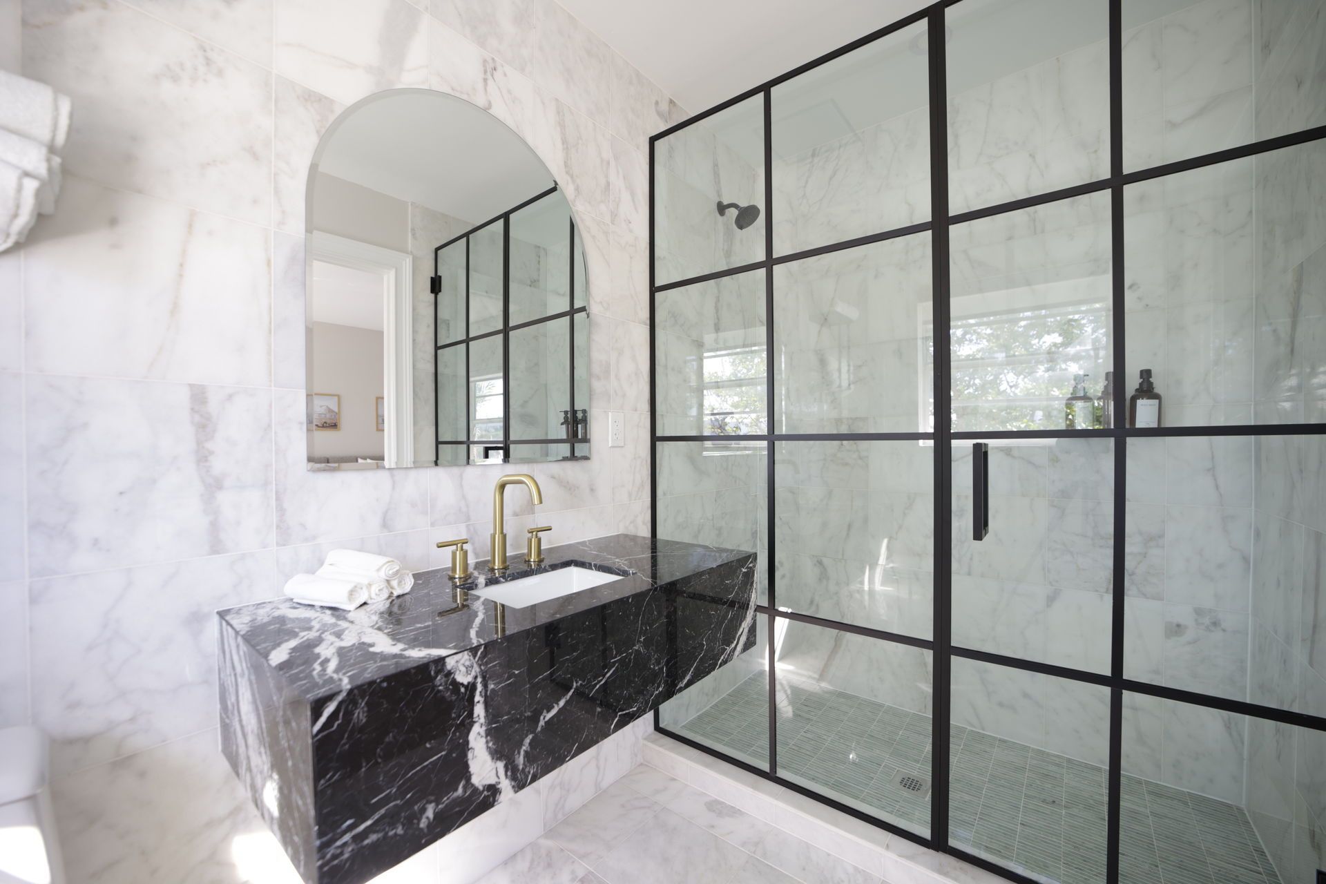 Elegant modern bathroom with marble walls and black marble vanity. Black-framed glass shower. Gold faucet. Arch mirror.