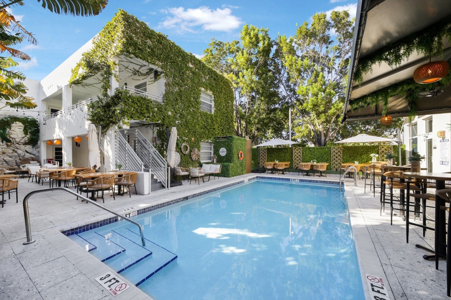 Poolside outdoor dining and swimming area at a hotel, with a vine-covered building.