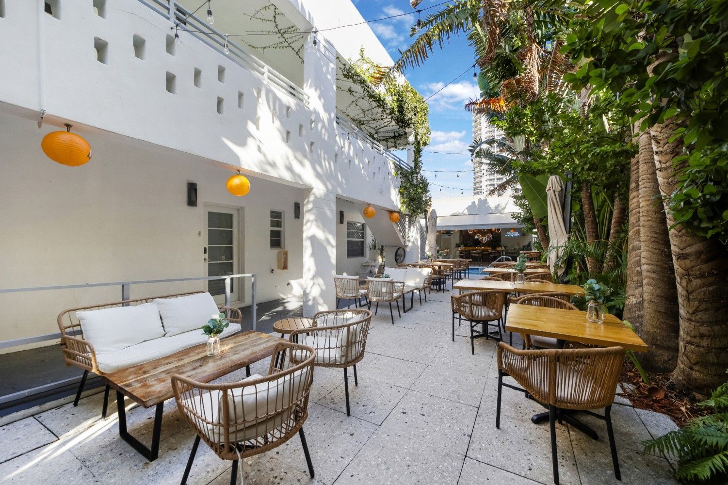 Outdoor patio restaurant with tables, chairs, and a couch. White walls, palm trees, and hanging lights.