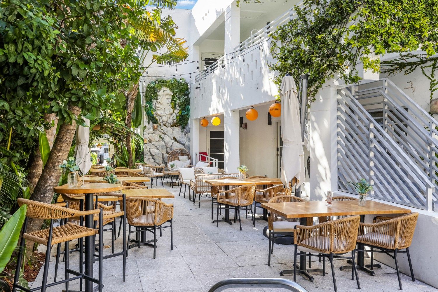 Outdoor restaurant with tables and chairs, under a white building with plants and umbrellas.