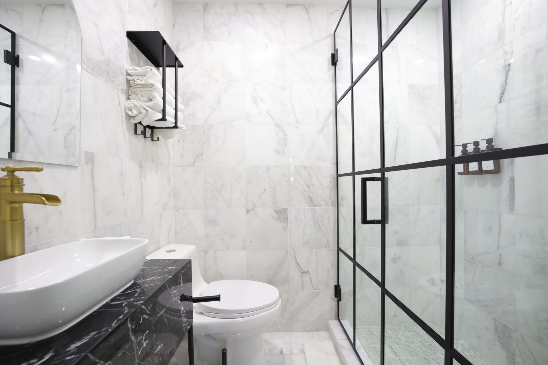 Modern bathroom with white marble tile, black framed shower, black shelf, and gold faucet.