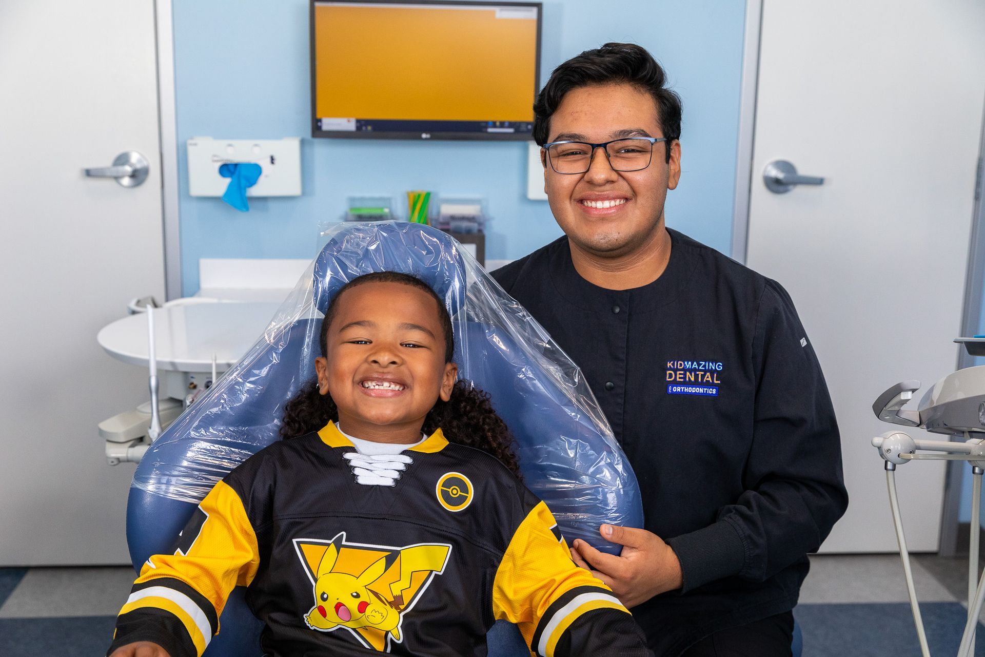 A smiling person in a navy scrub top stands beside a smiling child in a Pikachu jersey, both in a dental office.