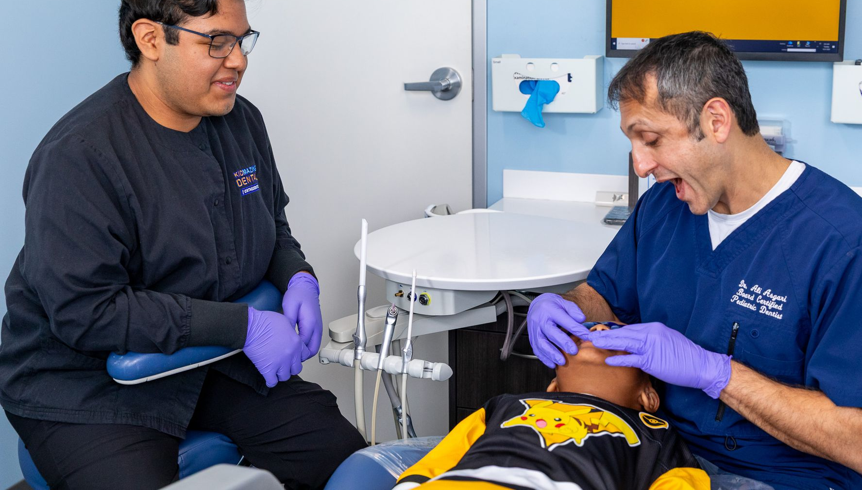 Dentists examine a child's teeth in a dental office. One assistant observes. They wear scrubs and gloves.