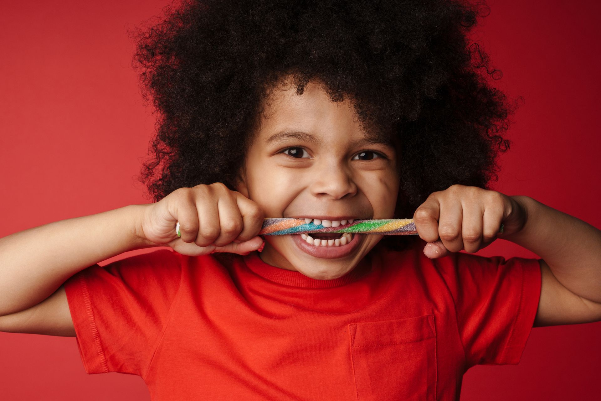 Young child with afro hair, smiling widely, biting on a colorful toothbrush against a red background.