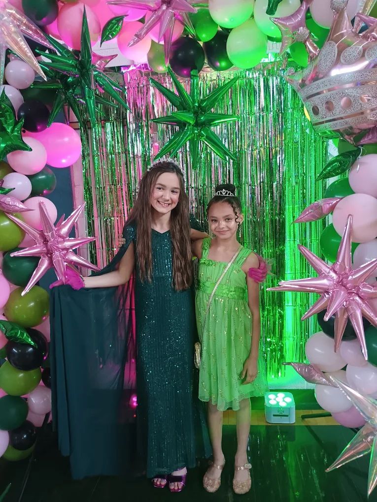 Two girls in formal dresses pose in front of a party backdrop with balloons and streamers.