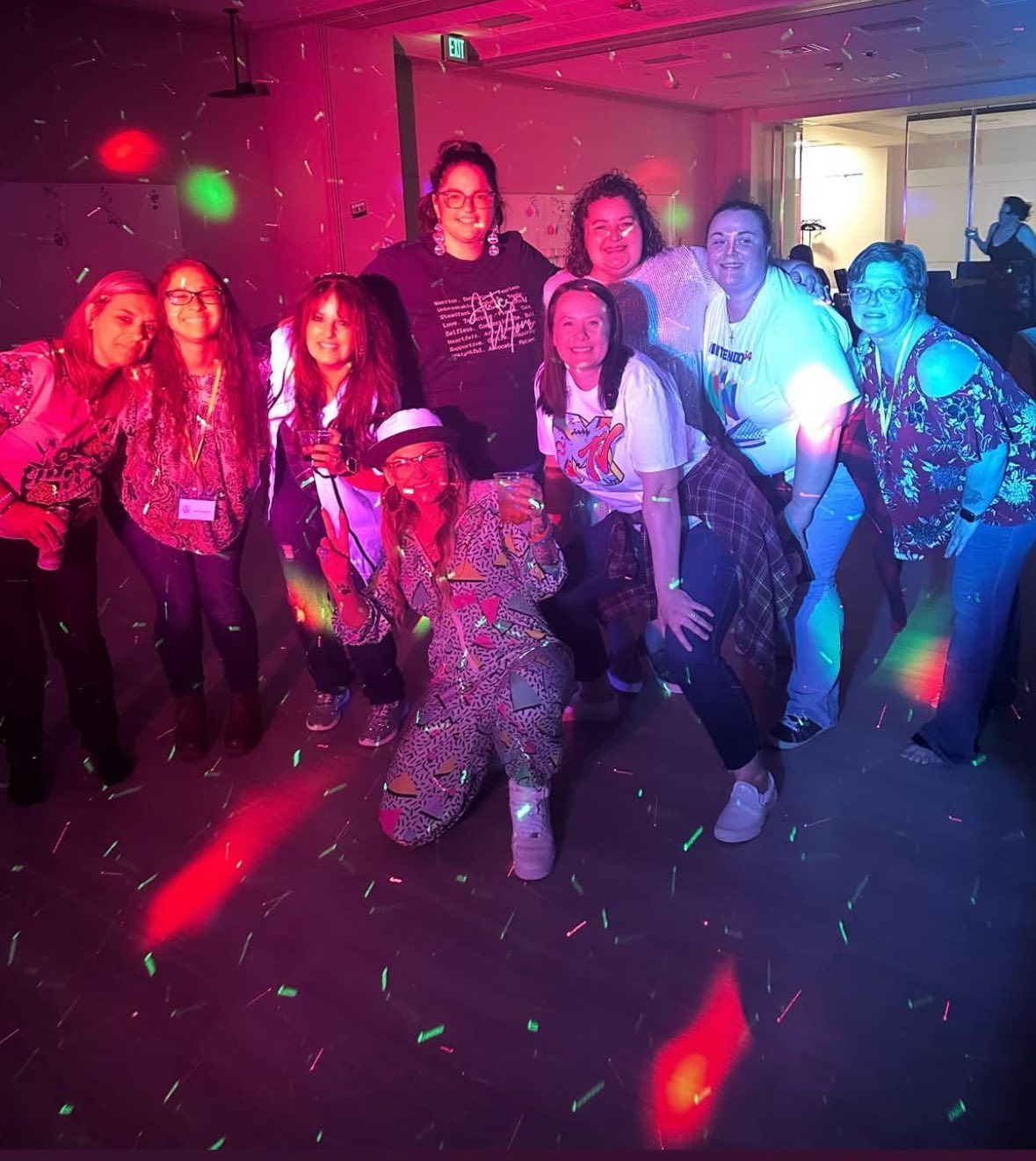 A group of nine people stand together at a party with colorful disco lights and confetti, smiling at the camera.