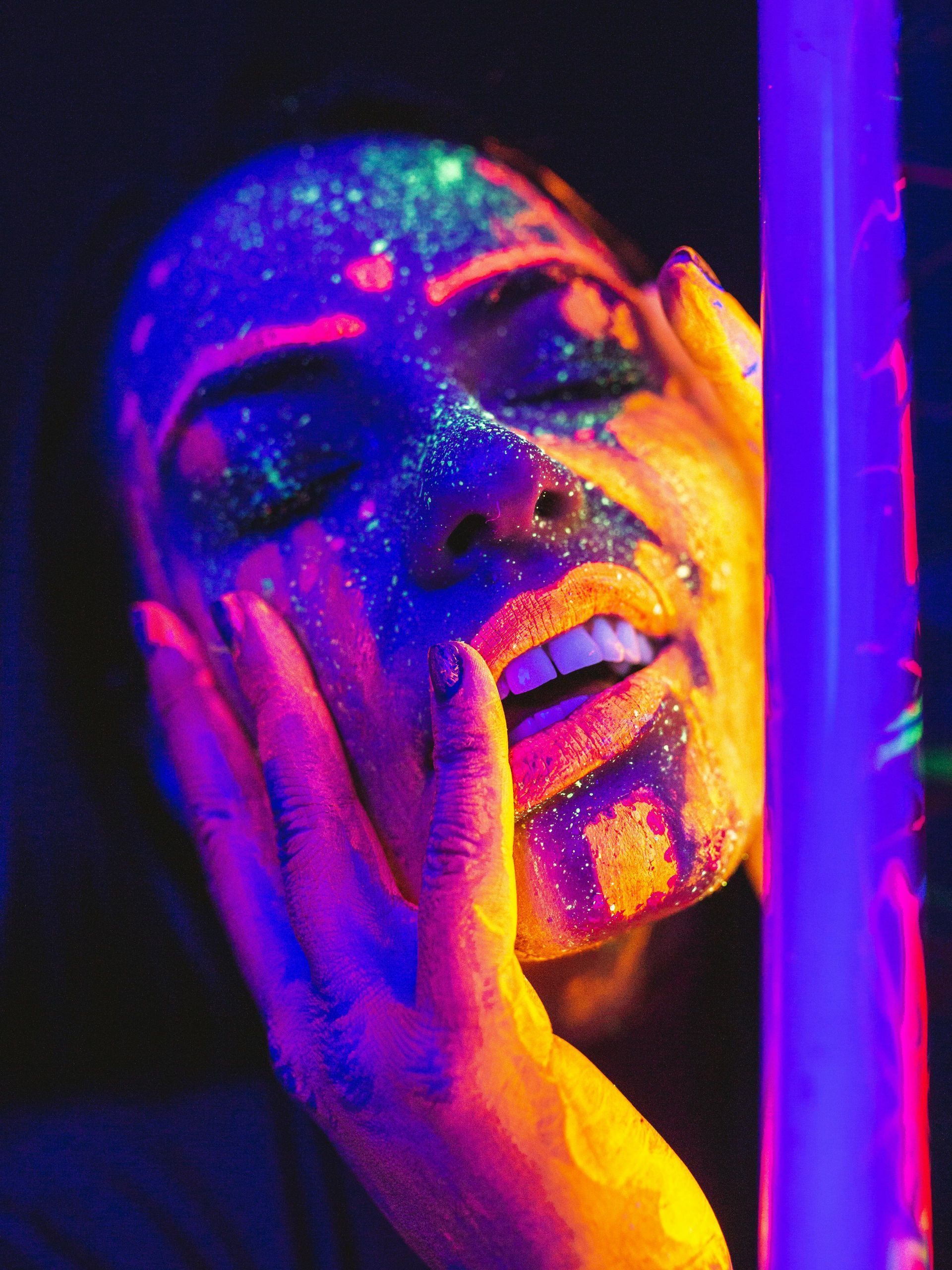 A face covered in glowing, neon ultraviolet face paint, with hands held to the side of the face in a dark, moody setting.