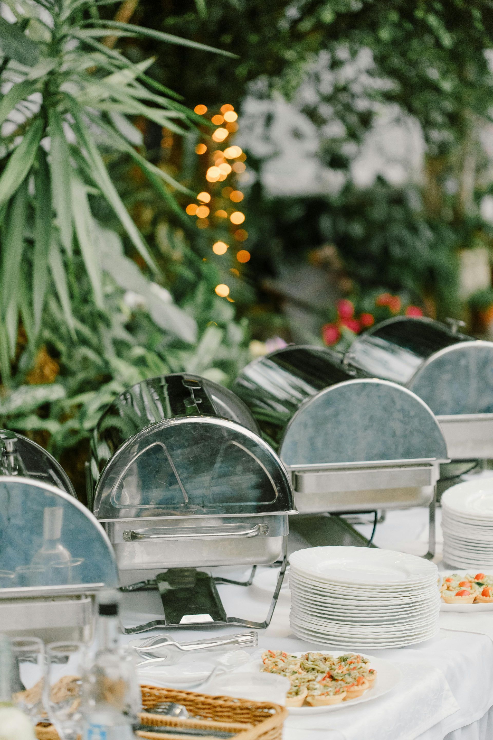 A buffet table outdoors with silver chafing dishes and stacks of white plates in a lush, green garden setting.