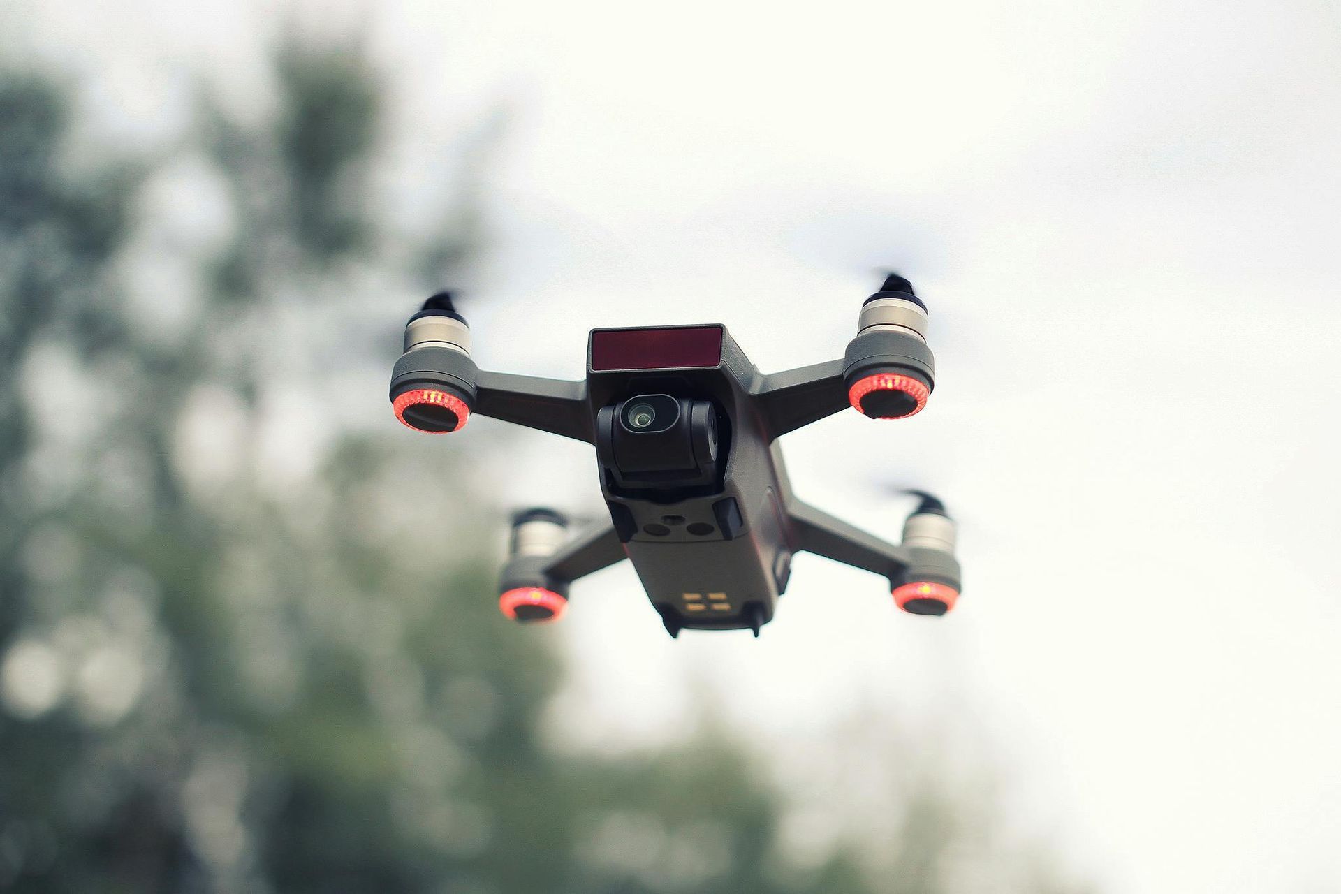 A dark grey quadcopter drone with orange-accented motor mounts hovering against a soft-focus, leafy green background.