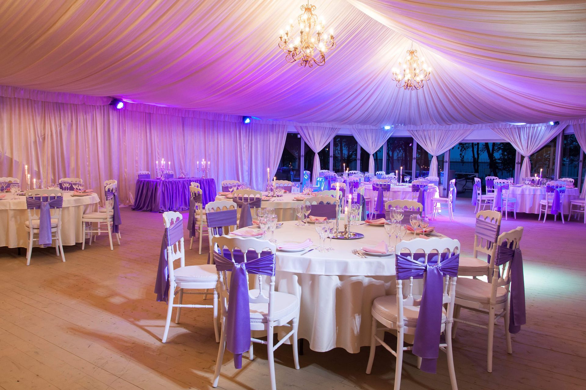 Elegant wedding reception hall with white tables and chairs, purple accents, and chandeliers.