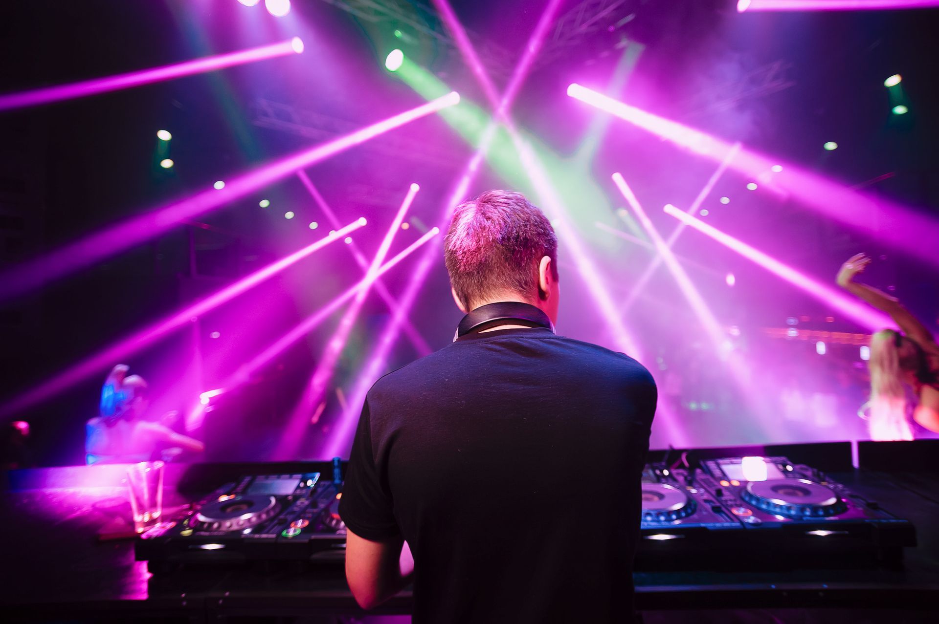 A DJ facing a dance floor under bright, vibrant pink and green stage lights in a nightclub setting.