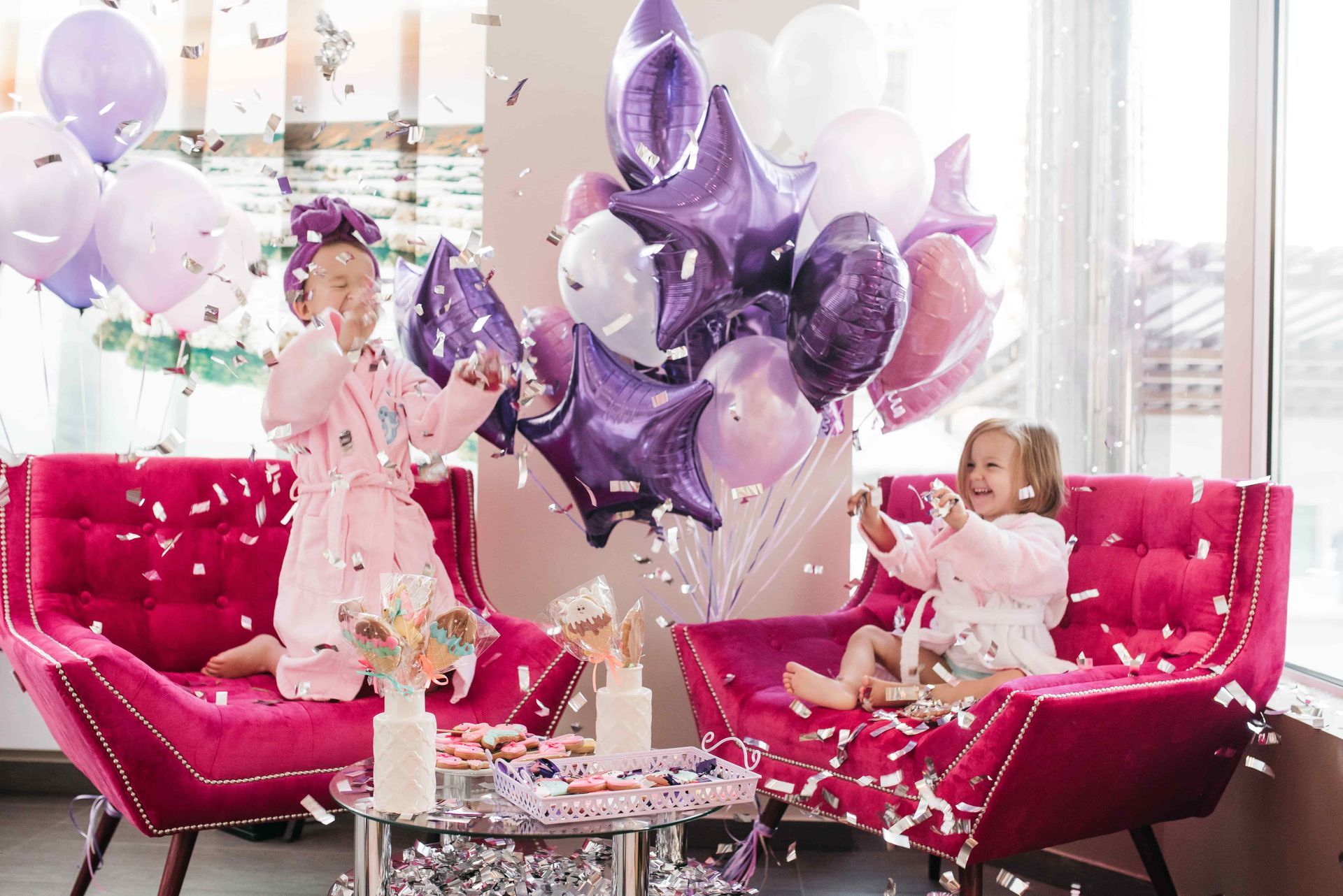 Two children in pink robes throw confetti near balloons in front of a window.