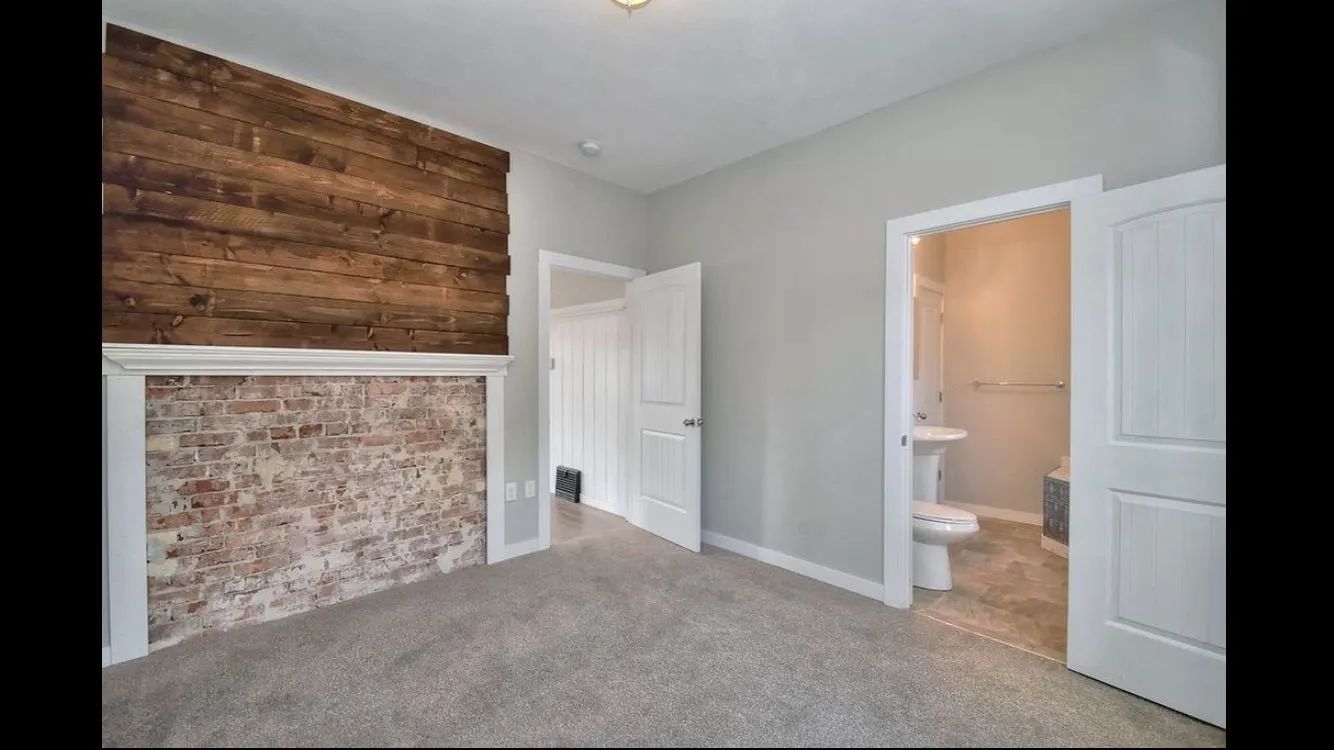 Empty bedroom with gray carpet, white doors, and a wood accent wall above a brick fireplace.
