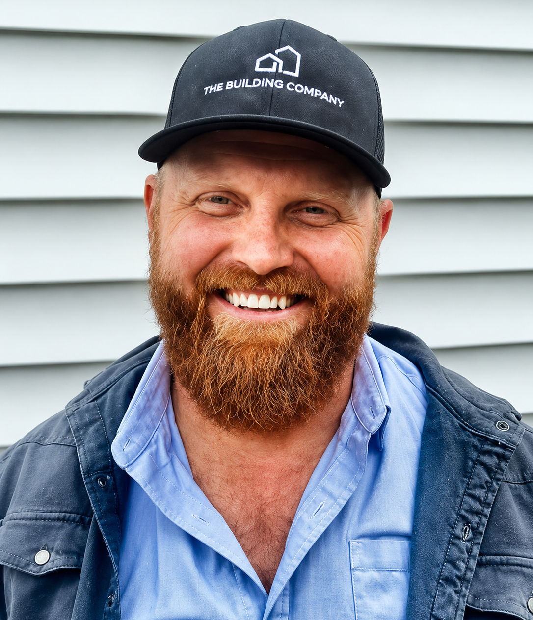 Smiling bearded man in a black cap and blue shirt standing against light gray siding