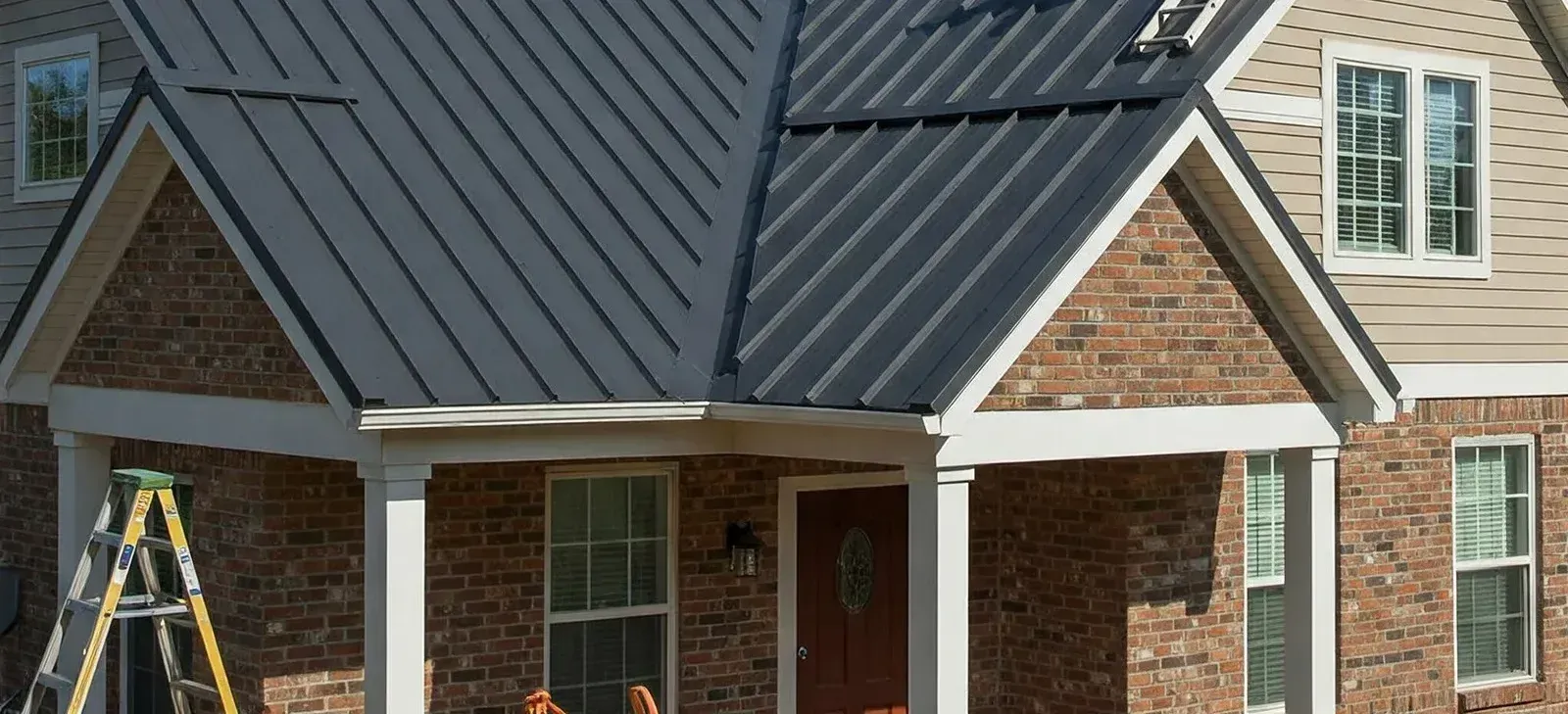 A house with a gray metal roof and red brick exterior, white trim around the windows, and a porch wi