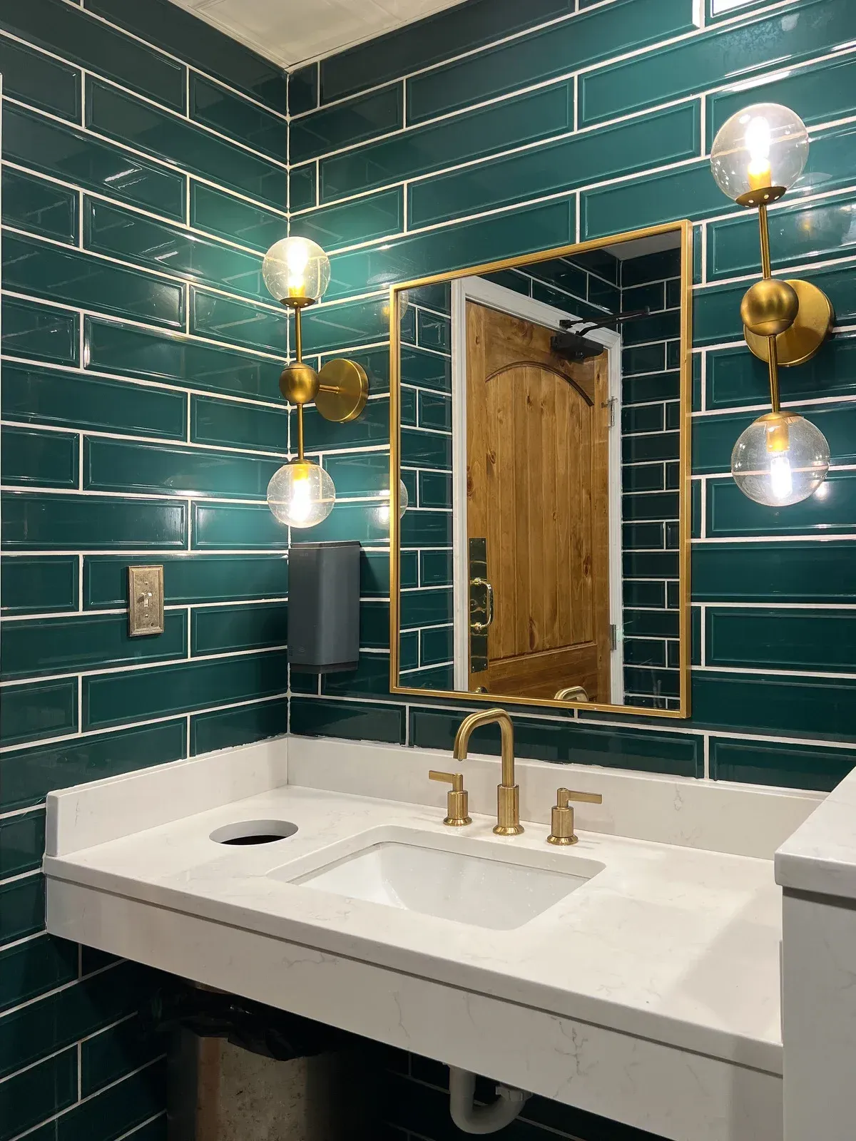 A bathroom with teal tile walls, a white countertop, gold fixtures, and a mirror reflecting a wooden door.