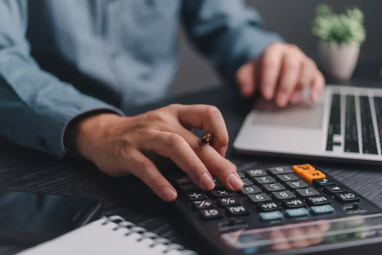A person is using a calculator in front of a laptop computer.