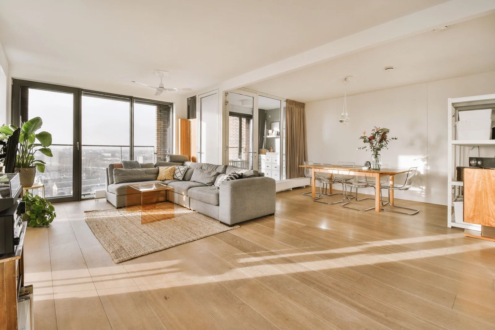 Bright open-plan living room with gray sofa, dining table, and large windows overlooking a balcony.